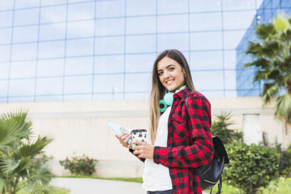 retrato-sonriente-estudiante-joven-que-sostiene-libros-taza-cafe-llevar-que-coloca-delante-edificio-universidad